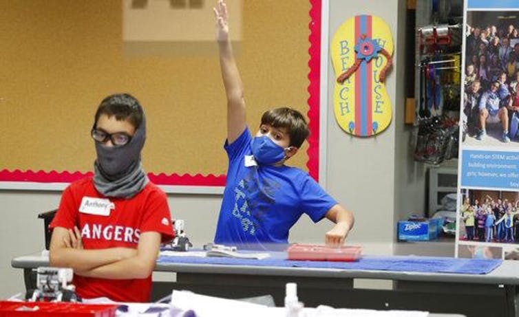 Two boys in a classroom wearing masks, with one raising his hand.