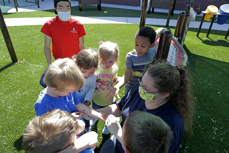 A group of kids without masks gather around a teacher wearing a mask outside.